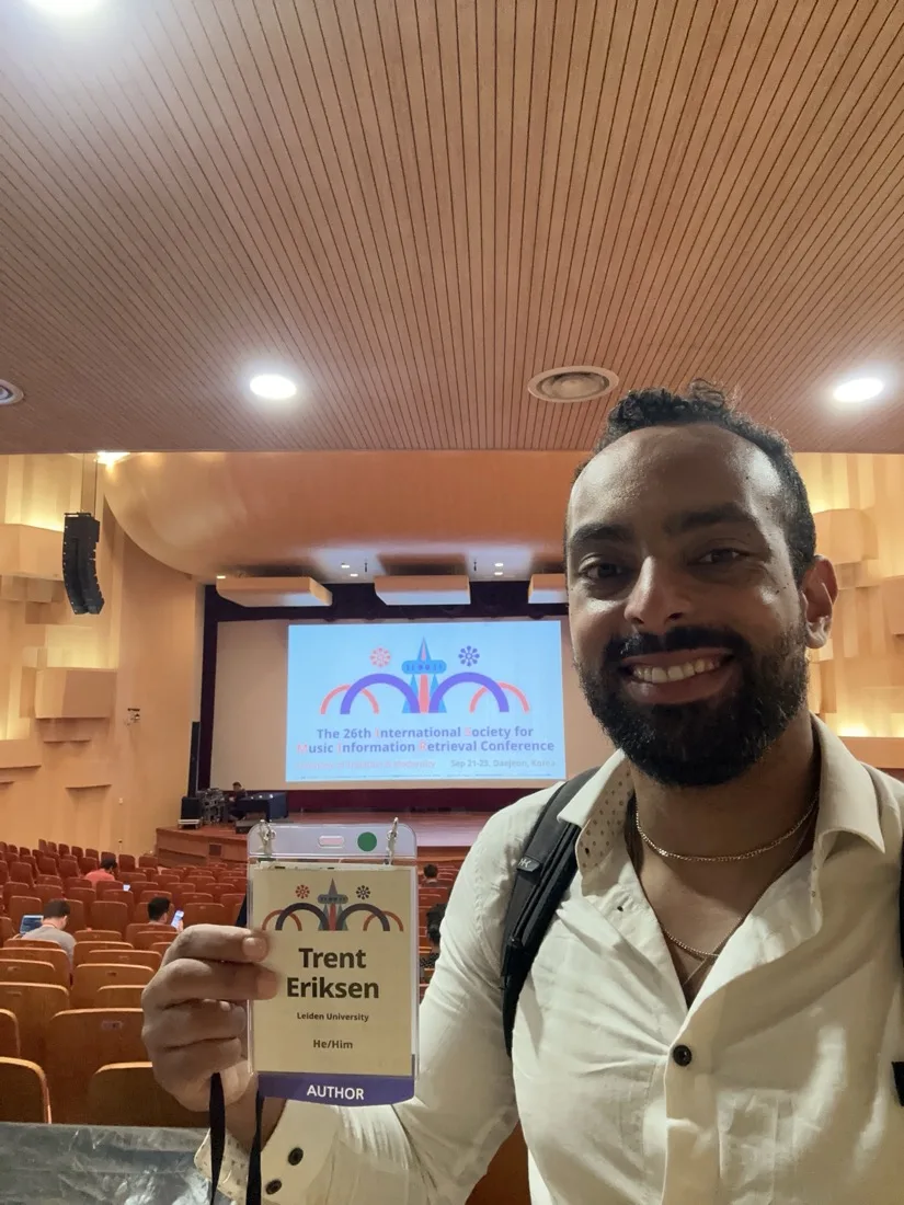 Trent Eriksen holding his ISMIR 2025 author badge in the conference hall, with the 26th ISMIR title screen behind him.